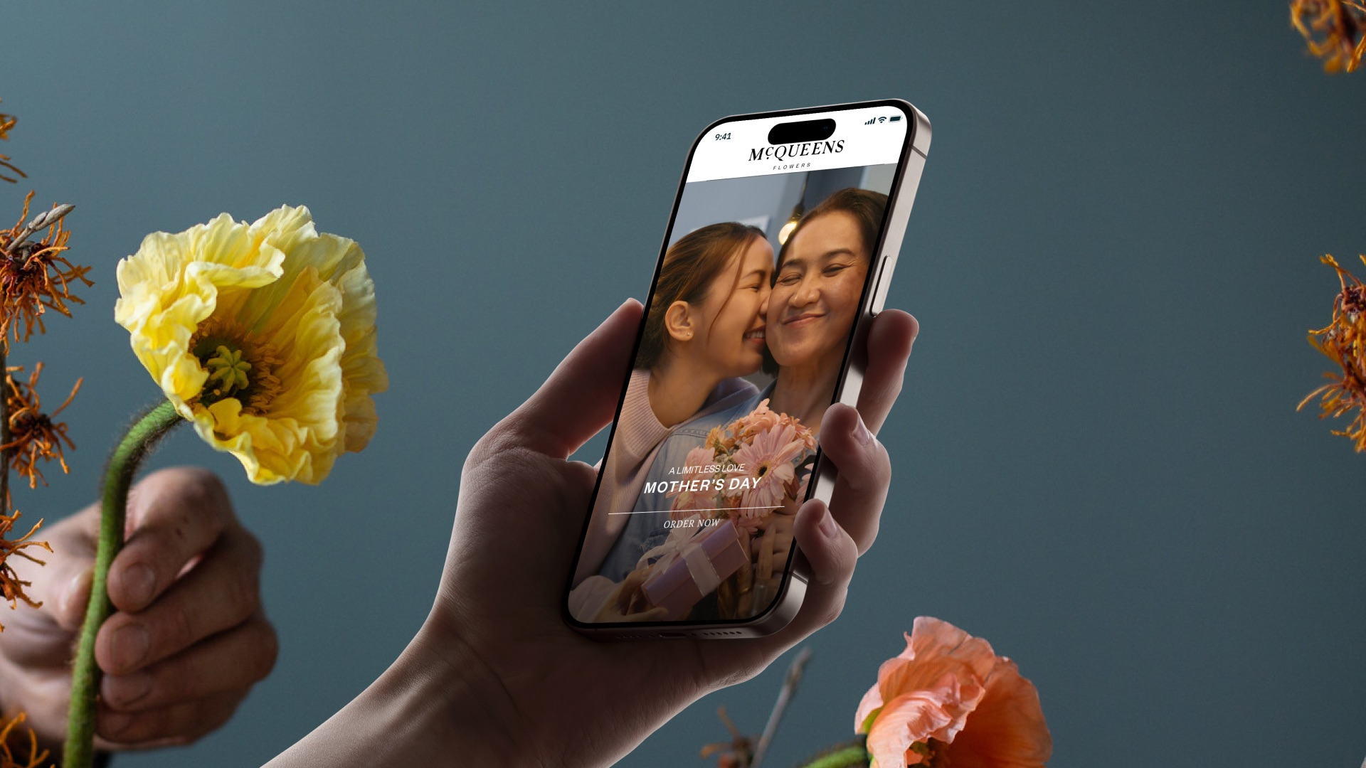 A hand holding an iPhone showing the McQueens home screen, a Mother's Day banner with a daughter kissing her mother, between yellow and coral poppies and dried witch hazel on a slate-blue wall