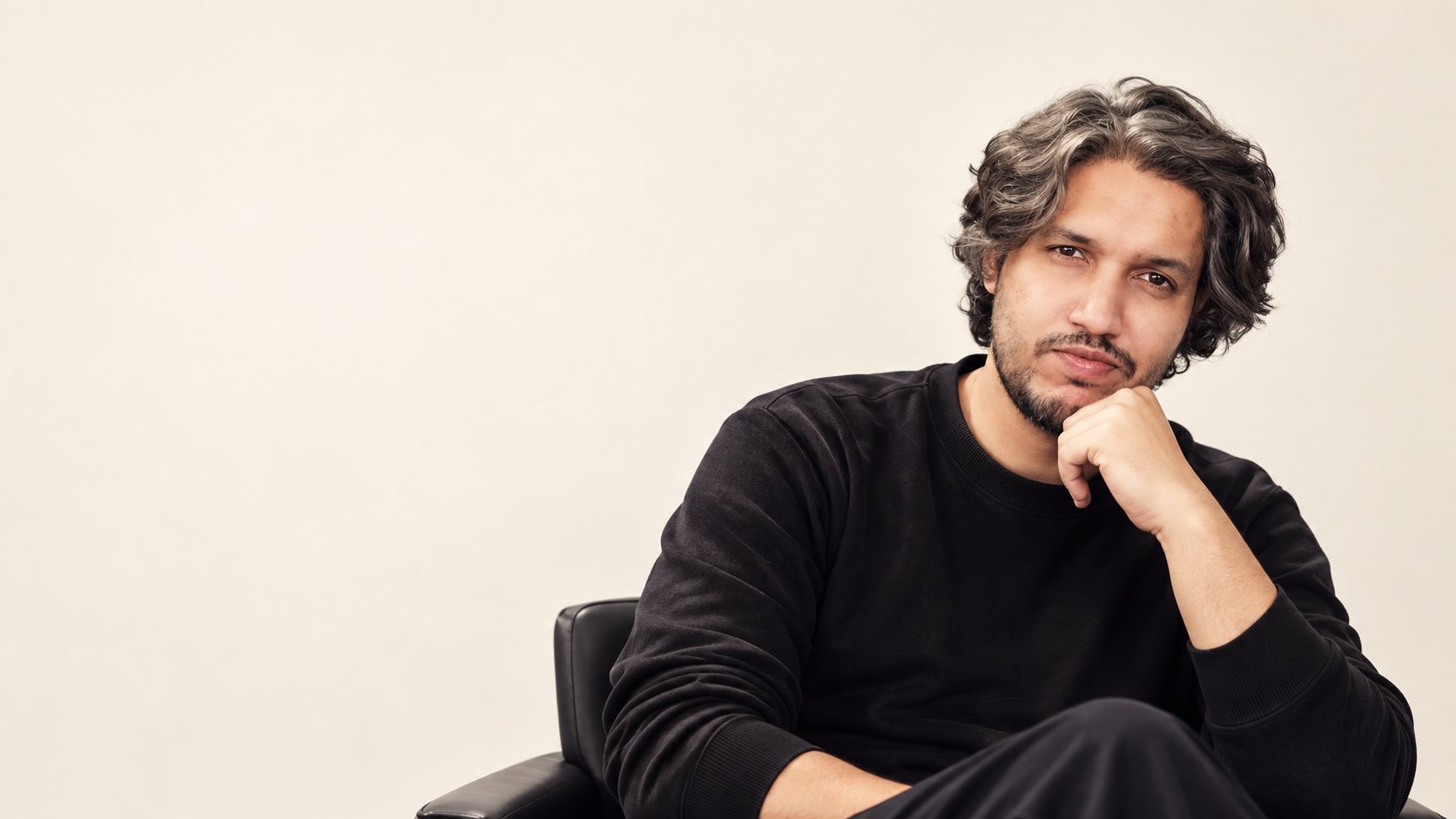 Zair Abbas, UX/UI designer based in Karachi, Pakistan, seated in a black chair against a plain cream backdrop, chin resting on his hand